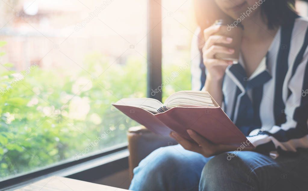 Focus on book. Relaxed Asian Woman holding a cup of coffee and r