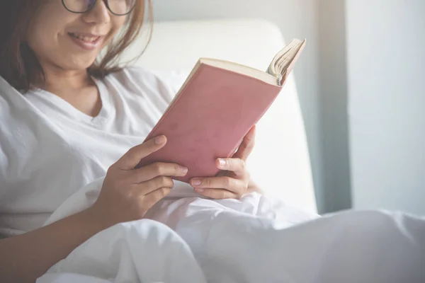 Frau mit asiatischer Brille liest ein Buch auf ihrem Bett. — Stockfoto