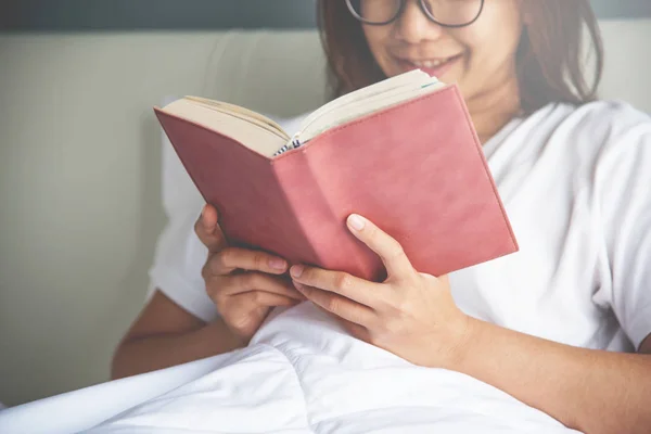 Frau mit asiatischer Brille liest ein Buch auf ihrem Bett. — Stockfoto