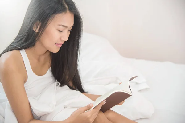 Relajada chica asiática leyendo un libro en su cama . — Foto de Stock