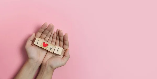 Manos sosteniendo cubos de madera con texto Love y corazón rojo en ba rosa — Foto de Stock