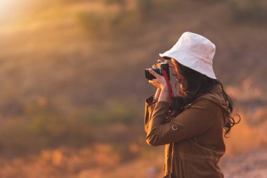 Güzel bir genç kadın yolcunun mutlu gülümseyen hayat tarzı portresi akşam vakti doğa arka planında fotoğraf çekerken eğleniyor. Güzel bir fotoğrafçı kız, dışarıda dolaşırken şapka takar.. 