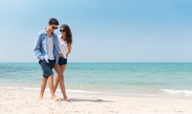 Romantik genç bir çiftin portresi günlük güneş gözlüğü takıyor ve mavi gökyüzüyle kumsalda yürüyor. Mutlu gülümseyen yakışıklı adam ve aşık güzel kadın. Sevgili, sevgili, aşk..