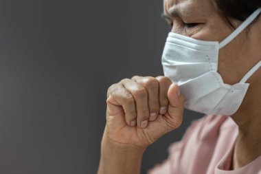 Asyalı yaşlı kadın beyaz cerrahi maske takıyor ve öksürüyor. Gri arka plan karantinası yüzünden stresli yaşlı kadın yüzü. Coronavirus veya Covid-19. Salgın, Salgın, Hijyenik, Sağlıklı ve Sağlık Hizmetleri konsepti. Sancak ve kopyalama alanı.