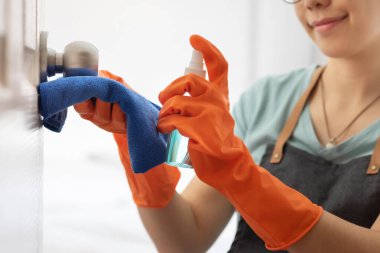 Turuncu lastik eldivenli çekici Asyalı kadın kapıyı siliyor, kapı tokmağında alkol temizleme spreyi var. Temizlik için dezenfeksiyon. Ev işleriyle gülümseyen güzel bir ev hanımı. Coronavirus, COVID-19 Önleme Konsepti.