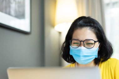 Mutlu gülümseyen Asyalı kadın Freelancer gözlük takıyor, eğleniyor ve karantina sırasında insanlarla konuşmak için dizüstü bilgisayarla görüntülü görüşme yapmaktan keyif alıyor. Sarı gömlekli genç bayan mavi cerrahi maske takıyor.