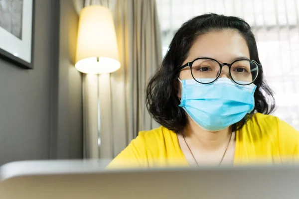 Happy Asian female wear eyeglasses using laptop computer during quarantine, isolated. Young Woman in yellow shirt wear blue surgical mask working from home during pandemic virus. Covid-19, Coronavirus, Healthy, Prevention, Protection.