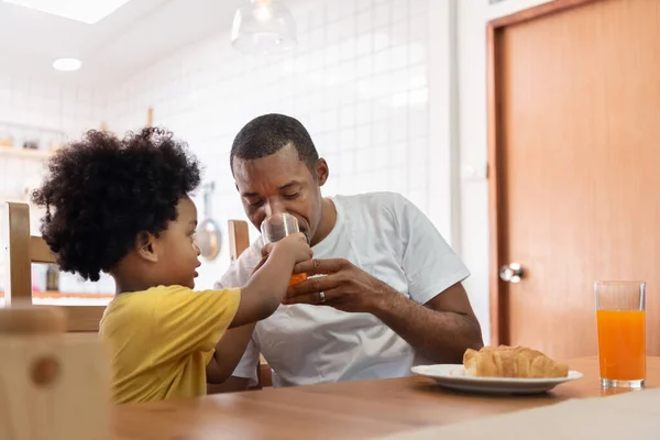 Happy African American family enjoying with drinking orange juice in dining room. Joyful Black Man in white cloth and little kid boy in yellow shirt with breakfast or brunch. Lifestyle.