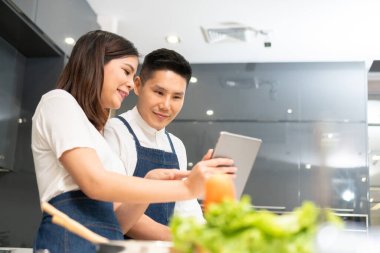 Çekici gülümseyen Asyalı çift evde yemek pişiriyor ve dijital tablet kullanıyorlar. Mutlu genç adam ve beyaz elbiseli ve mavi önlüklü güzel kadın salata için sebze hazırlıyorlar.