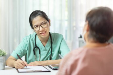 Yeşil üniformalı güzel Asyalı kadın doktor gözlük takıyor kağıda bilgi yazıyor ve hastanedeki yaşlı kadın hastaya tavsiye veriyor. Hastalık, Covid 19, Coronavirus
