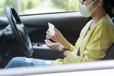 Yeşil ya da sarı tişörtlü Asyalı kadın araba sürmeden önce Coronavirus ya da Covid 19 'u önlemek için avuçlarına dezenfektan jel sürerken koruyucu maske takıyor. Temizlik, antiseptik, hijyenik, sağlıklı.