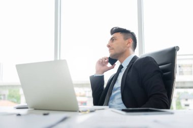Siyah takım elbiseli yakışıklı iş adamı, iş yerinde iş yerinde dizüstü bilgisayar üzerinde çalışırken ortaklar ve müşterilerle iş görüşmeleri yapmak için cep telefonu kullanıyor..
