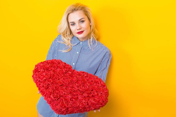 Beautiful young woman in blue shirt holding big red heart made of roses on yellow background