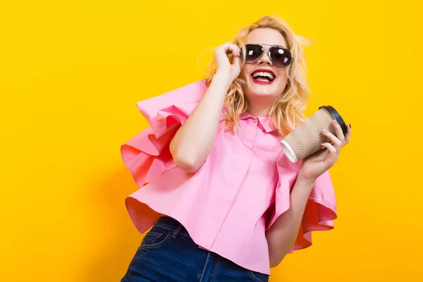 stylish young woman in pink blouse posing with paper cup of coffee on yellow background
