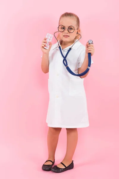 可爱的小女孩在医疗制服与听诊器和药片在粉红色背景 — 图库照片