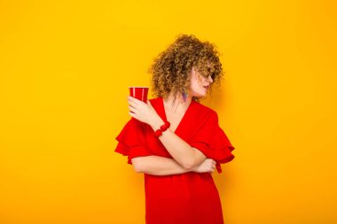 Çekici genç kıvırcık saçlı kadın kırmızı elbise ve plastik bardak içki ile tutarak güneş gözlüğü 