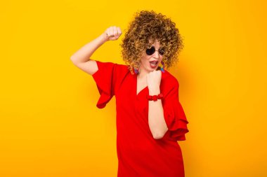 Çekici genç kıvırcık saçlı kadın kırmızı elbise ve sarı arka planı poz güneş gözlüğü 