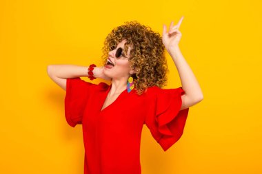 Çekici genç kıvırcık saçlı kadın kırmızı elbise ve zafer jest gösterilen güneş gözlüğü