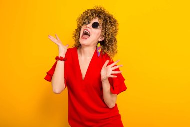 Heyecanlı genç kıvırcık saçlı kadın kırmızı elbise ve Sarı zemin üzerine güneş gözlüğü