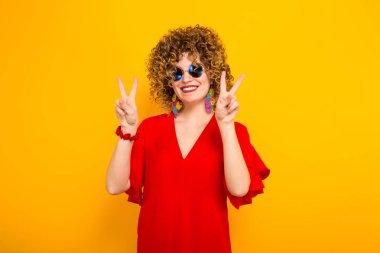 Çekici genç kıvırcık saçlı kadın kırmızı elbise ve zafer jest gösterilen güneş gözlüğü
