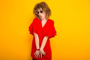 Komik genç kıvırcık saçlı kadın kırmızı elbise ve dilini gösteren güneş gözlüğü