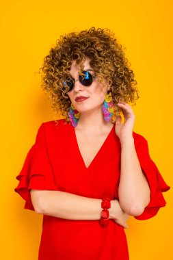 Çekici genç kıvırcık saçlı kadın kırmızı elbise ve sarı arka planı poz güneş gözlüğü 