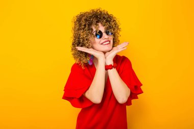 Çekici genç kıvırcık saçlı kadın kırmızı elbise ve çenesinin altında ellerini tutarak ve gülümseyen güneş gözlüğü