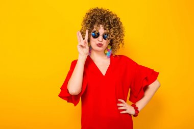Çekici genç kıvırcık saçlı kadın kırmızı elbise ve zafer jest gösterilen güneş gözlüğü