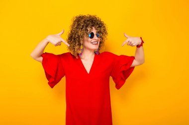 Çekici genç kıvırcık saçlı kadın kırmızı elbise ve parmakları ile işaret güneş gözlüğü 