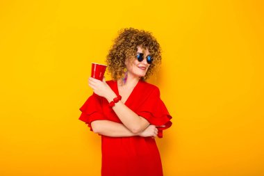 Çekici genç kıvırcık saçlı kadın kırmızı elbise ve plastik bardak içki ile tutarak güneş gözlüğü 