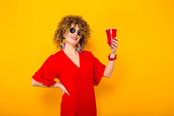 Привлекательная молодая кудрявая женщина в красном платье и солнцезащитных очках с пластиковой чашкой с напитком
 