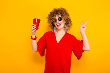 Çekici genç kıvırcık saçlı kadın kırmızı elbise ve plastik bardak içki ile tutarak güneş gözlüğü 