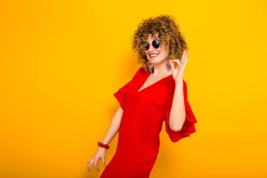 Çekici genç kıvırcık saçlı kadın kırmızı elbise ve ok jest gösterilen güneş gözlüğü 