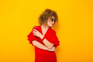 Çekici genç kıvırcık saçlı kadın kırmızı elbise ve plastik bardak içki ile tutarak güneş gözlüğü 