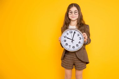 Duvar saati tutarak ve kamera sarı arka plana karşı gülümseyen sevimli küçük esmer kız 
