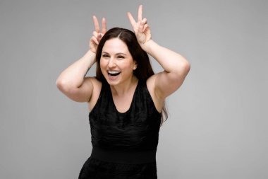 Studio gri duvar farklı ifadeler gösterilen basit siyah gömlekli uzun lüks saçlı güzel curvy esmer kadın. Gülüyor ve boynuzları yapıyor.