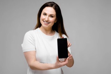 Farklı ifadeler gri duvar ve gösteren boş ekran smartphone üzerinde gösterilen basit beyaz t-shirt uzun lüks saçlı güzel curvy esmer kadın