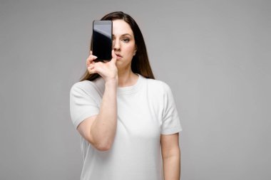 Onun smartphone ile poz Studio gri duvar farklı ifadeler gösterilen basit beyaz t-shirt uzun lüks saçlı güzel curvy esmer kadın.