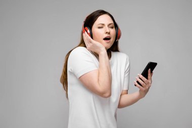 Basit beyaz t-shirt enjoing müzik ile akıllı telefon ve kulaklık, uzun süre lüks saçlı güzel curvy esmer kadın Studio gri duvar farklı ifadeler gösterilen.