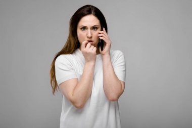 Telefonda konuşurken gri duvara farklı ifadeler gösterilen basit beyaz t-shirt uzun lüks saçlı güzel curvy esmer kadın.