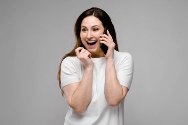 Telefonda konuşurken gri duvara farklı ifadeler gösterilen basit beyaz t-shirt uzun lüks saçlı güzel curvy esmer kadın.