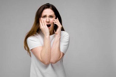 Telefonda konuşurken gri duvara farklı ifadeler gösterilen basit beyaz t-shirt uzun lüks saçlı güzel curvy esmer kadın.