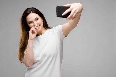 Farklı ifadeler Studio gri duvar selfie onun Smartphone için poz sırasında gösterilen basit beyaz t-shirt uzun lüks saçlı güzel curvy esmer kadın.