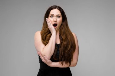 Studio gri duvar farklı ifadeler gösterilen basit siyah gömlekli uzun lüks saçlı güzel curvy esmer kadın. Şok ve sürpriz.