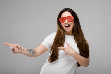 Basit beyaz t-shirt ve gri duvar Studio farklı ifadeler gösterilen kırmızı plastik parti güneş gözlüğü uzun lüks saçlı güzel curvy esmer kadın.