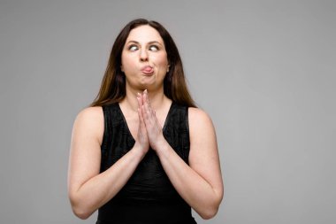 Studio gri duvar farklı ifadeler gösterilen basit siyah gömlekli uzun lüks saçlı güzel curvy esmer kadın. Dua eden eller ve komik yüz buruşturma.