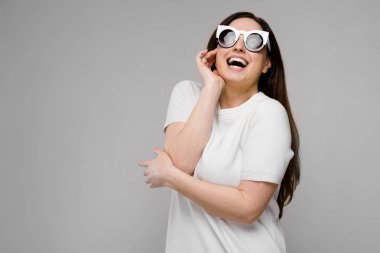 Studio gri duvar farklı ifadeler gösterilen basit Beyaz tişört ve şık güneş gözlüğü uzun lüks saçlı güzel curvy esmer kadın.