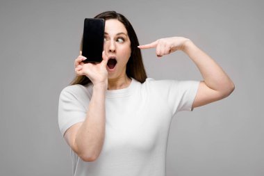 Onun smartphone ile poz Studio gri duvar farklı ifadeler gösterilen basit beyaz t-shirt uzun lüks saçlı güzel curvy esmer kadın.