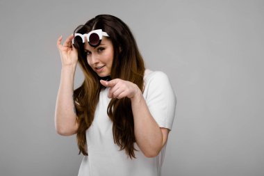 Studio gri duvar farklı ifadeler gösterilen basit Beyaz tişört ve şık güneş gözlüğü uzun lüks saçlı güzel curvy esmer kadın.