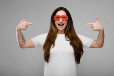 Basit beyaz t-shirt ve gri duvar Studio farklı ifadeler gösterilen kırmızı plastik parti güneş gözlüğü uzun lüks saçlı güzel curvy esmer kadın.